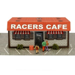 May include: A miniature model of a red and white brick building with a sign that reads "Racers Cafe". The building has a red roof and a white awning. There are three miniature people standing in front of the building.