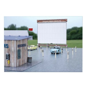 May include: A model drive-in cinema scene featuring a large screen with the text "SUGAR CREEK DRIVE IN". There's a refreshment stand, a light blue convertible, and a yellow car. The scene is set on a grey surface.
