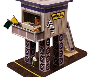 May include: A miniature race control tower model with the word "WEIAND" in yellow. The tower is gray with blue trim, supported by blue and silver supports. Small figures stand on the base and inside the tower. Checkered and colored flags are attached.
