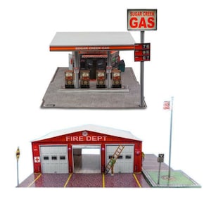 Op de afbeelding: Een miniatuurmodelset met een Sugar Creek Gas-tankstation met vier brandstofpompen en een hoog prijsbord, naast een rood Fire Dept-gebouw met witte garagedeuren, een ladder en een vlaggenmast, alles op gedetailleerde basisplatforms.
