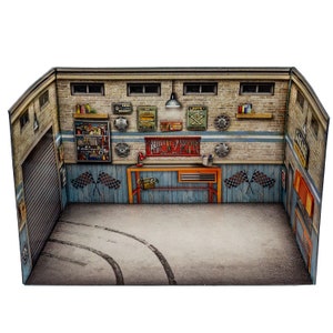 May include: A miniature cardboard model of a garage with a workbench, tools, and tire-related signs. The garage has a gray concrete floor with tire tracks and a gray metal garage door.