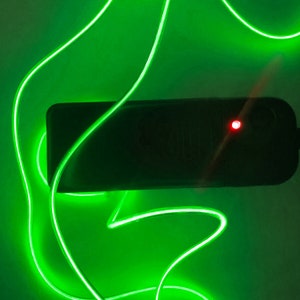 May include: A black rectangular device with a red light on the side, connected to a green neon light cord.