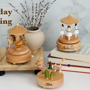 May include: Three wooden music boxes featuring a carousel with horses, a carousel with ballerinas, and a seesaw with small figures. The carousel with ballerinas is engraved with the name Amelia, and the seesaw base is engraved with the name Chloe.
