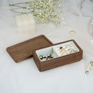 May include: A wooden jewelry box with a hinged lid, divided into two compartments. One compartment holds rings and the other holds earrings. The box is made of dark wood with a natural grain.