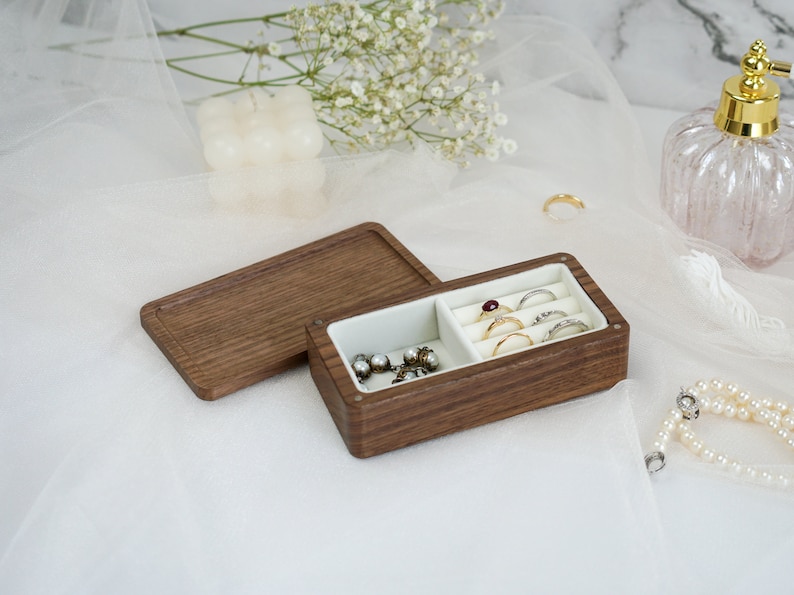 May include: A wooden jewelry box with a hinged lid, divided into two compartments. One compartment holds rings and the other holds earrings. The box is made of dark wood with a natural grain.
