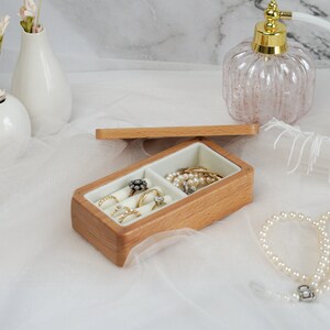 May include: A wooden ring box with two compartments. One compartment is filled with rings and the other with a pearl necklace. The box is open and sitting on a white surface.