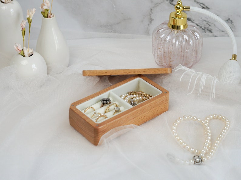 May include: A wooden ring box with two compartments. One compartment is filled with rings and the other with a pearl necklace. The box is open and sitting on a white surface.