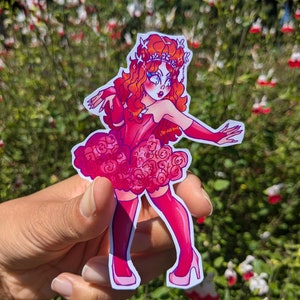 May include: A sticker featuring a stylized illustration of a woman with red hair and a pink dress. The woman is wearing a crown and high heels. The sticker is held in front of a background of green foliage and red flowers.