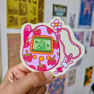 May include: A pink and green sticker featuring a strawberry-themed design. The sticker depicts a retro handheld game with a screen showing a cartoon character. The design includes strawberries, a flower, a bunny charm, and a beaded chain.