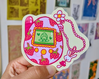 May include: A pink and green sticker featuring a strawberry-themed design. The sticker depicts a retro handheld game with a screen showing a cartoon character. The design includes strawberries, a flower, a bunny charm, and a beaded chain.