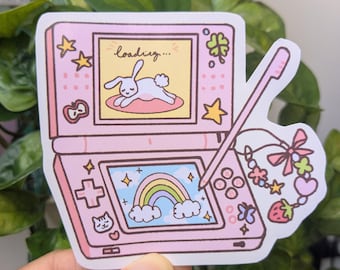 May include: A pink handheld gaming console sticker featuring a top screen with a rabbit and the text loading... and a bottom screen displaying a rainbow. A stylus is attached by a beaded charm with a pink bow, a heart, and a strawberry.