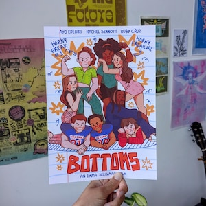 May include: A poster featuring a colorful illustration of a group of people. The poster has the word "BOTTOMS" in large red letters. The names of the actors are listed at the top. The poster is held by a hand.