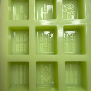 May include: A green silicone mold with six square cavities. Each cavity has a different design, including a flower, a series of lines, and a letter "W" with a letter "M" inside.