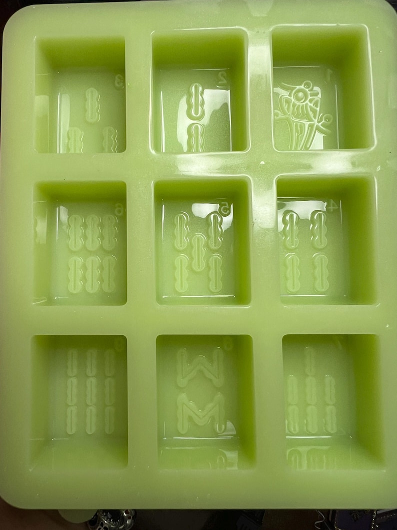 May include: A green silicone mold with six square cavities. Each cavity has a different design, including a flower, a series of lines, and a letter "W" with a letter "M" inside.