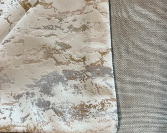 May include: Close-up of two fabric swatches. The top swatch is a cream-colored fabric with a subtle marbled pattern in shades of gold, gray, and white. The bottom swatch is a plain, light gray fabric with a woven texture.