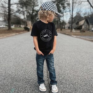 May include: A young boy wearing a black t-shirt with a white graphic that says "Long Live Boyhood" and a black and white checkered baseball cap. He is wearing blue jeans and white sneakers.
