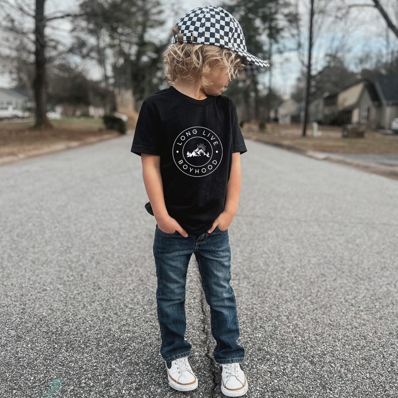 May include: A young boy wearing a black t-shirt with a white graphic that says "Long Live Boyhood" and a black and white checkered baseball cap. He is wearing blue jeans and white sneakers.