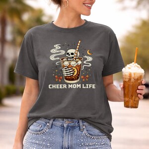 May include: Dark grey t-shirt featuring a skull graphic in a drink with a straw and the text "CHEER MOM LIFE". The shirt is worn with light blue jeans. The person is holding a large iced coffee.