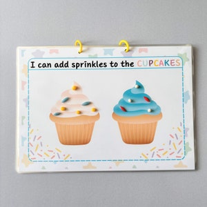 May include: A children's learning activity book with two yellow binder rings. The page features two illustrated cupcakes with pink and blue frosting, and the text "I can add sprinkles to the CUPCAKES". The page is decorated with colorful sprinkles and star shapes.