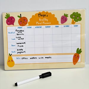 Puede incluir: Un planificador de comidas amarillo y blanco con el texto "Maya's Healthy Meal Planner". El planificador tiene una cuadr&iacute;cula para las comidas diarias, con ilustraciones de frutas y verduras. Un marcador blanco y negro est&aacute; delante del planificador.