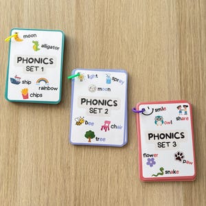 May include: Three brightly coloured phonics flashcards with illustrations and words. The cards are labelled "PHONICS SET 1", "PHONICS SET 2", and "PHONICS SET 3". Each card has a different coloured border and includes words like "moon", "ship", "bee", "owl", and "flower".