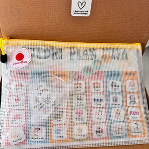Op de afbeelding: Een witte mesh-ritszak met een gele rits en een gekleurde bedrukte kaart erin. De kaart heeft een raster van gekleurde vierkanten met afbeeldingen en tekst. De tekst op de kaart zegt "EDNI PLAN XTRA". De tas heeft een klein rood insectenlogo met de tekst "LearnBug" erop.