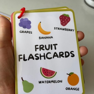 Pode incluir: Um conjunto de cart&otilde;es de mem&oacute;ria de frutas com ilustra&ccedil;&otilde;es de uvas, banana, morango, fatia de melancia, pera e laranja. O t&iacute;tulo "FRUIT FLASHCARDS" est&aacute; impresso em negrito. Os cart&otilde;es s&atilde;o presos com um clip vermelho.