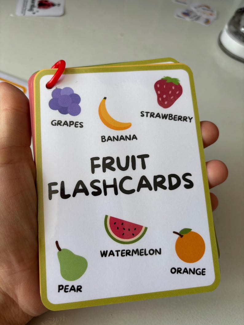 Pode incluir: Um conjunto de cart&otilde;es de mem&oacute;ria de frutas com ilustra&ccedil;&otilde;es de uvas, banana, morango, fatia de melancia, pera e laranja. O t&iacute;tulo "FRUIT FLASHCARDS" est&aacute; impresso em negrito. Os cart&otilde;es s&atilde;o presos com um clip vermelho.