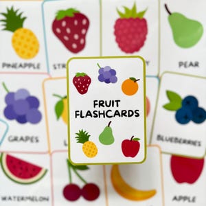 Pode incluir: Cart&otilde;es de mem&oacute;ria de frutas com ilustra&ccedil;&otilde;es coloridas. O cart&atilde;o central exibe o texto "FRUIT FLASHCARDS" e apresenta um morango, uvas, laranja, abacaxi, pera e ma&ccedil;&atilde;.