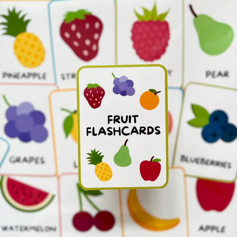Pode incluir: Cart&otilde;es de mem&oacute;ria de frutas com ilustra&ccedil;&otilde;es coloridas. O cart&atilde;o central exibe o texto "FRUIT FLASHCARDS" e apresenta um morango, uvas, laranja, abacaxi, pera e ma&ccedil;&atilde;.