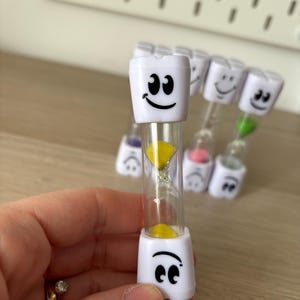 May include: A clear hourglass with white plastic end caps featuring a smiling face design. The hourglass contains yellow sand. Several similar hourglasses are visible in the background.