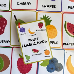 Pode incluir: Um conjunto de cart&otilde;es de mem&oacute;ria de frutas coloridas com ilustra&ccedil;&otilde;es e r&oacute;tulos. O cart&atilde;o central diz "FRUIT FLASHCARDS" e mostra imagens de morango, banana, melancia e laranja. Outros cart&otilde;es mostram cerejas, uvas e mirtilos.