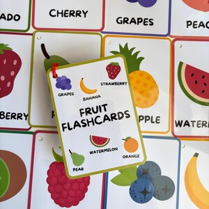 Pode incluir: Um conjunto de cart&otilde;es de mem&oacute;ria de frutas coloridas com ilustra&ccedil;&otilde;es e etiquetas. O cart&atilde;o central diz "FRUIT FLASHCARDS" e exibe imagens de melancia, laranja e pera. Outros cart&otilde;es mostram cerejas, uvas, morangos e mirtilos.