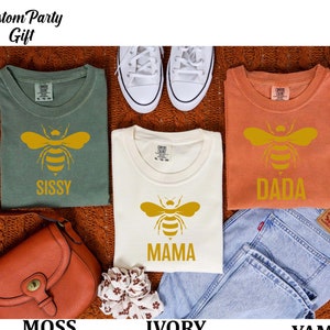 May include: Three t-shirts with a bee graphic and the words "Sissy", "Mama", and "Dada" printed on them. The shirts are in the colors moss, ivory, and yam.