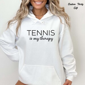A woman wearing a white hoodie with the text "TENNIS is my therapy" printed on it.