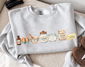 May include: A light gray crewneck sweatshirt featuring a graphic print of baking ingredients including a bottle of vanilla, a carton of eggs, a mixing bowl with a spoon, a jar labeled sugar, a measuring cup, a bag of flour, and a stick of butter.