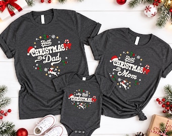 May include: Two dark gray short-sleeve t-shirts and one dark gray short-sleeve baby bodysuit. The shirts feature white text reading First Christmas as Dad and First Christmas as Mom, while the bodysuit reads My First Christmas with reindeer.