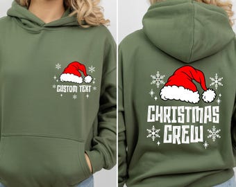 May include: An olive green hooded sweatshirt featuring a red and white Santa hat graphic with white snowflakes on the front chest above the text CUSTOM TEXT and on the back above the white text CHRISTMAS CREW in a bold, blocky font style.