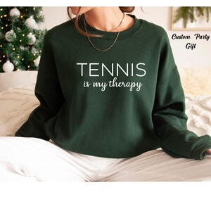 A person wearing a green sweatshirt with the text "TENNIS is my therapy" printed on it.