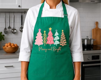 May include: A dark green apron featuring a graphic print of five stylized Christmas trees in shades of pink, yellow, and light blue. The text Merry and Bright is printed in a gold script font below the trees. The apron includes two front pockets.