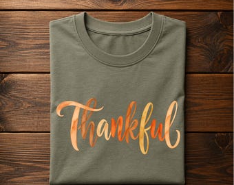 May include: A military green crew neck t-shirt featuring the word Thankful printed in a stylized orange and yellow watercolor script font across the center. The text H Military Green is visible in the top left corner of the image.
