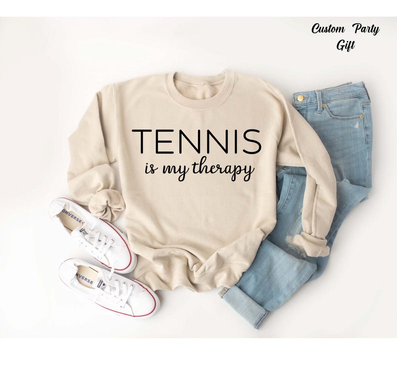 A beige sweatshirt with the text "TENNIS is my therapy" printed on it, along with a pair of blue jeans and white sneakers.