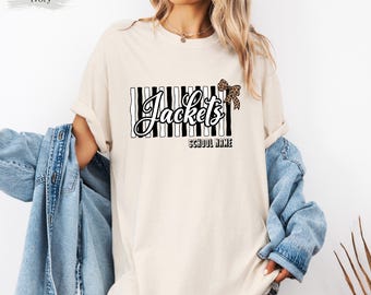 May include: An ivory short-sleeve t-shirt featuring a graphic with vertical black and white stripes, the word "Jackets" in a cursive script, a leopard print bow, and the text "SCHOOL NAME" printed in a bold black sans-serif font.
