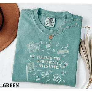 A turquoise t-shirt with a printed message, "HOWVER YOU COMMUNICATE, I AM LISTENING", along with various icons and symbols. The t-shirt is placed on a white surface, and there is a brown hat and some dried leaves nearby.