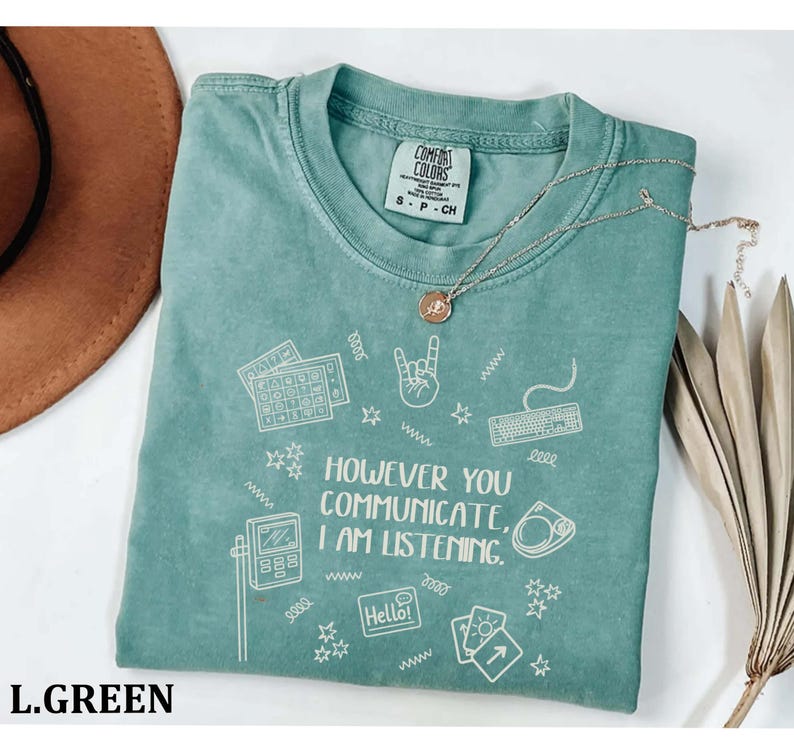 A turquoise t-shirt with a printed message, "HOWVER YOU COMMUNICATE, I AM LISTENING", along with various icons and symbols. The t-shirt is placed on a white surface, and there is a brown hat and some dried leaves nearby.