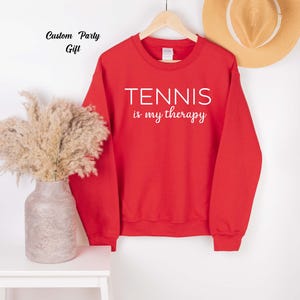 A red sweatshirt with the text "TENNIS is my therapy" printed on it, hanging on a wooden hanger. Next to the sweatshirt is a vase with dried flowers, and a straw hat is also visible in the background.