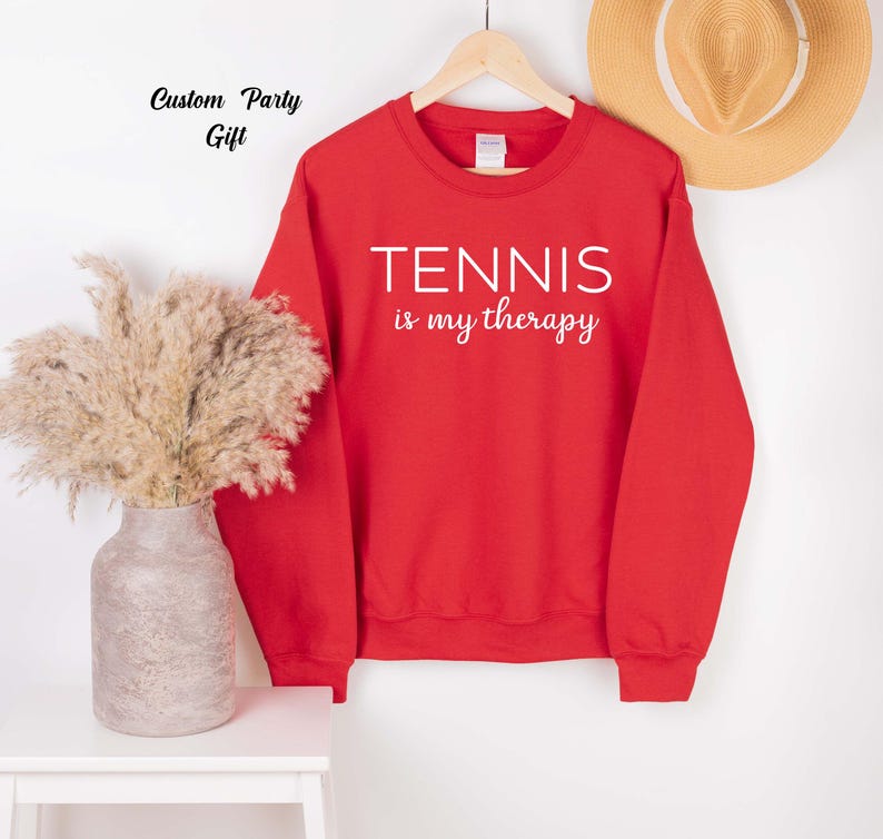 A red sweatshirt with the text "TENNIS is my therapy" printed on it, hanging on a wooden hanger. Next to the sweatshirt is a vase with dried flowers, and a straw hat is also visible in the background.