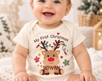 May include: A cream-colored short-sleeve baby bodysuit featuring a graphic of a reindeer with a red nose, colorful Christmas lights, and stars. The text reads My First Christmas and the name ROWAN in brown lettering on the front of the garment.