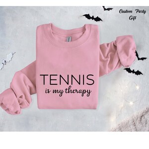 A pink sweatshirt with the text "TENNIS is my therapy" printed on it, along with bats scattered around the sweatshirt.