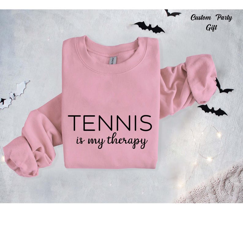 A pink sweatshirt with the text "TENNIS is my therapy" printed on it, along with bats scattered around the sweatshirt.